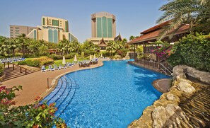 A large swimming pool with lounge chairs and umbrellas, surrounded by palm trees and a hotel building in the background. - Gulf Executive Residence (Manama)