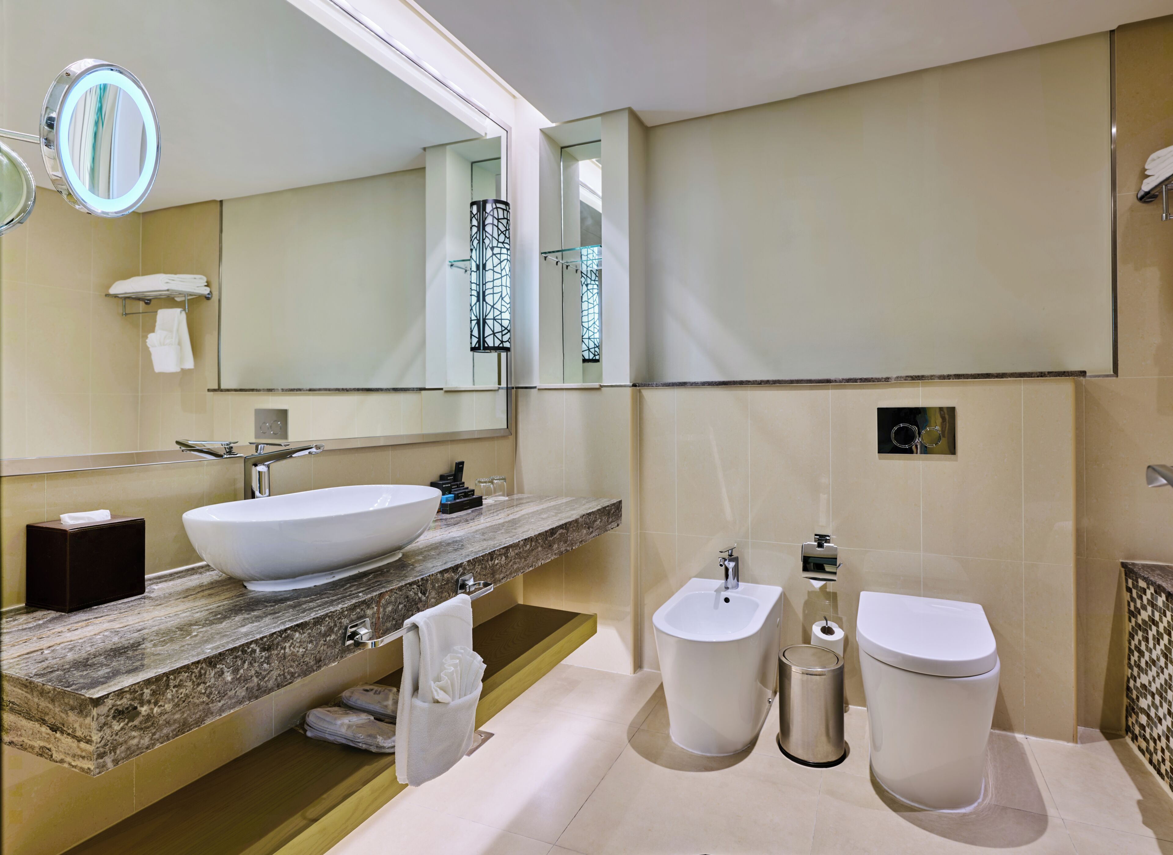 A modern bathroom with a large sink, bidet, and toilet, marble countertop, and a mirror with a light.