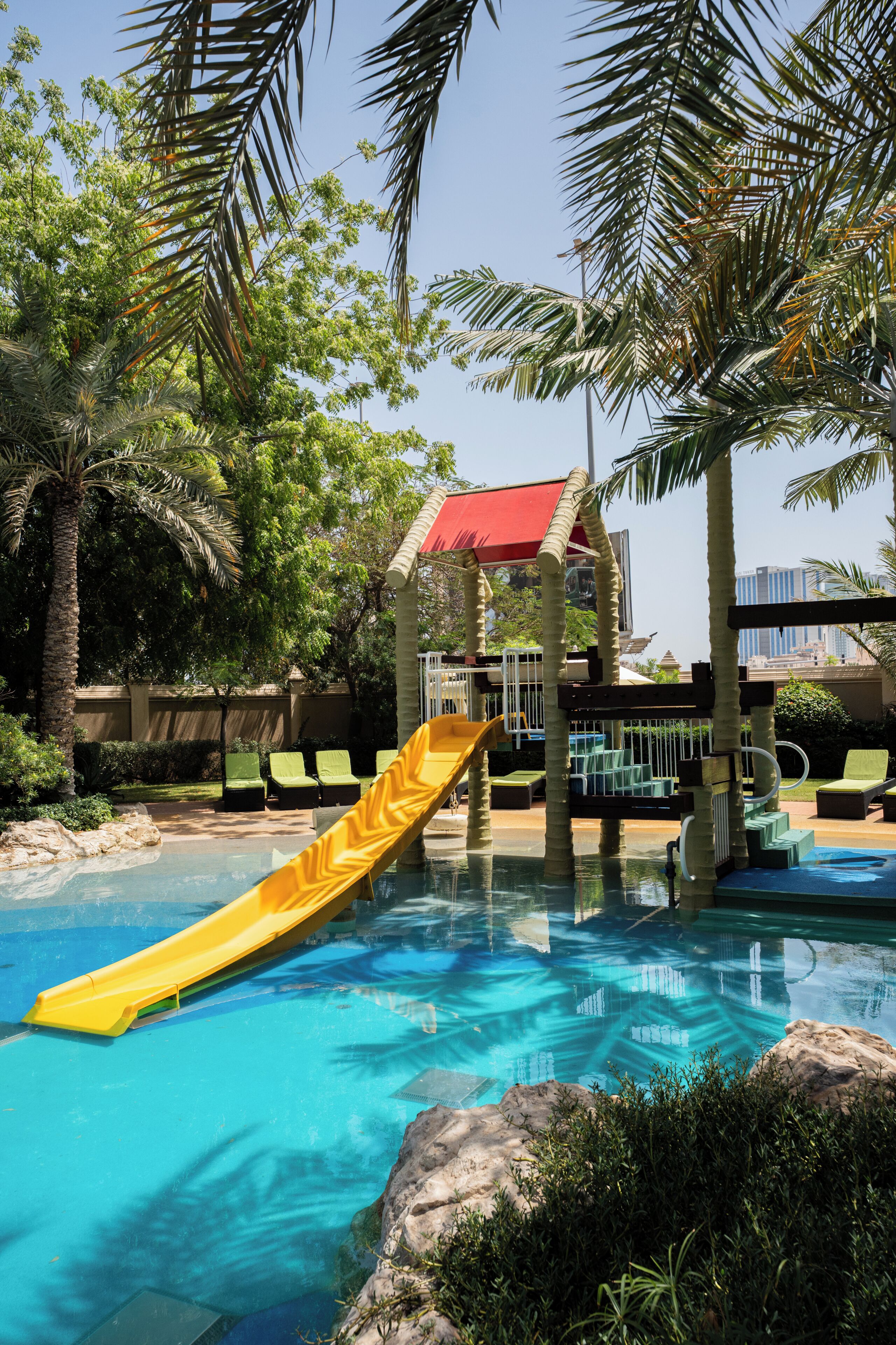 A pool area with a yellow slide, palm trees, and a red-roofed structure.