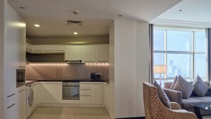 A modern kitchen with white cabinets, a built-in oven, and a washing machine. There is a seating area with a sofa and chairs near a large window. - Gulf Executive Residence (Manama)