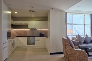 A modern kitchen with white cabinets, a built-in oven, and a washing machine. There is a seating area with a sofa and chairs near a large window. - Gulf Executive Residence (Manama)