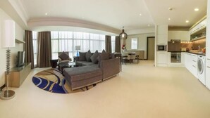 A modern living room with a sofa, a coffee table, and a TV. There is a dining area with a table and chairs. The room has large windows with curtains. - Gulf Executive Residence (Manama)