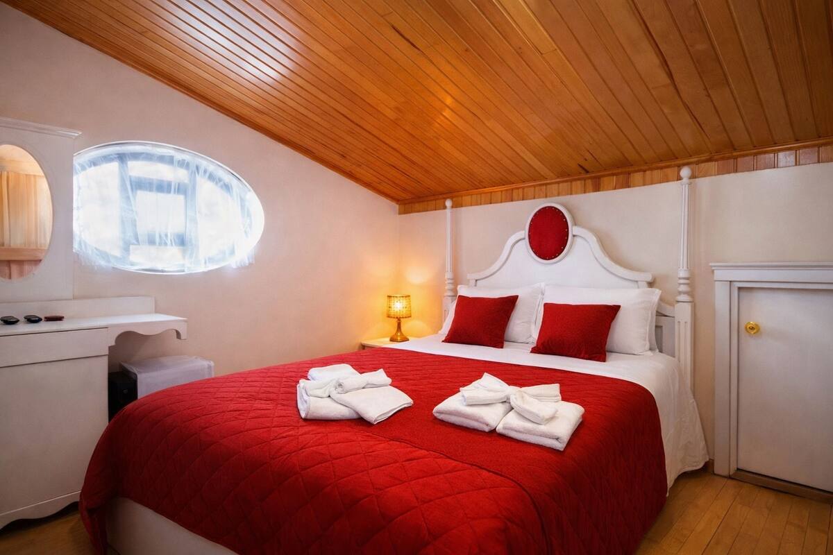 Une chambre d’hôtel avec un plafond en bois, un lit avec une literie rouge, une table de chevet avec une lampe, une armoire et une fenêtre donnant sur un paysage.