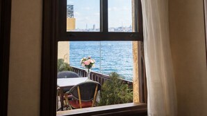 Ventana con vista al mar y al paisaje urbano, una silla y una mesa en el exterior.