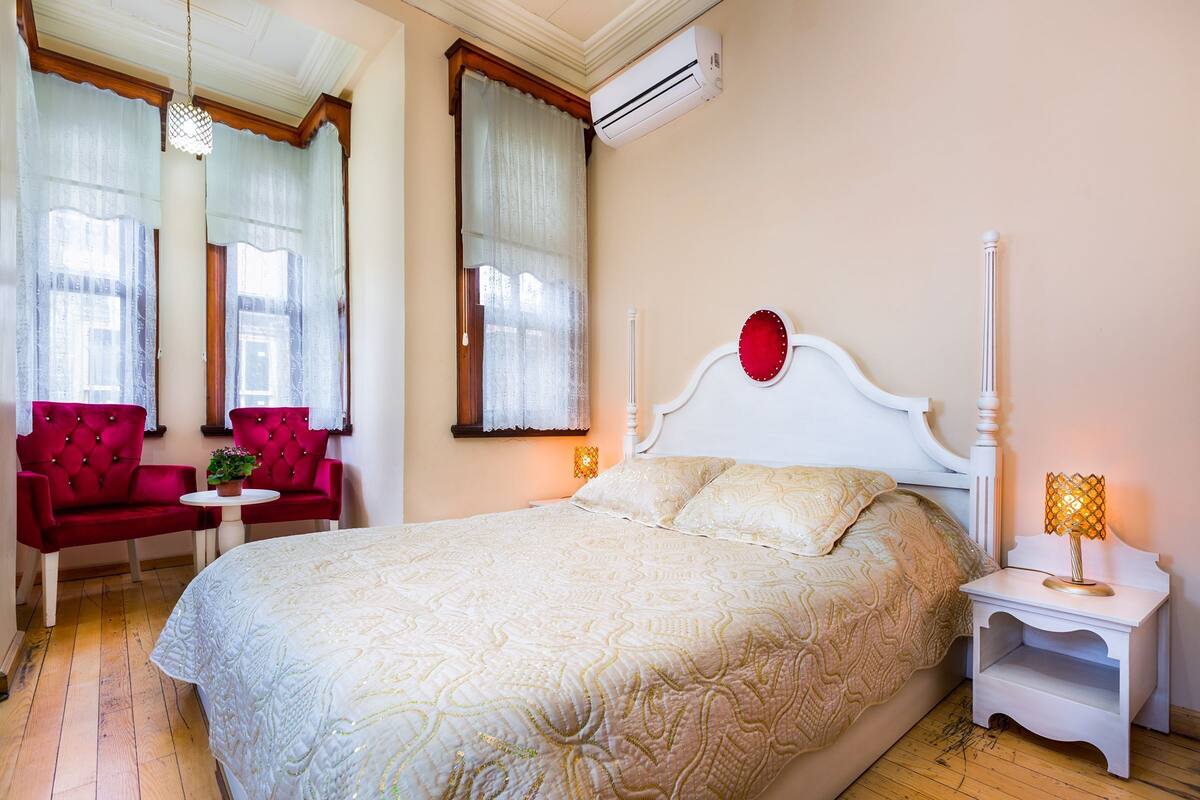 Une chambre à coucher avec un grand lit, une table de chevet, un fauteuil rouge et une fenêtre avec des rideaux.