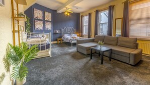 A living room with a sofa, coffee table, and a bed with a headboard. There is a ceiling fan, a window with curtains, and a plant.
