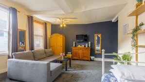 A living room with a grey sofa, a small table, a wooden cabinet, a TV, and a bed with floral bedding.