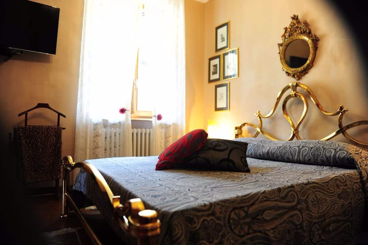 Una habitación de hotel con cama, televisor montado en la pared, radiador y ventana con cortinas.