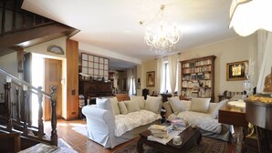 Ampla sala de estar com um sofá branco grande, uma mesa de centro e um candelabro. Há uma estante cheia de livros e uma escada de madeira que leva a outro cômodo.