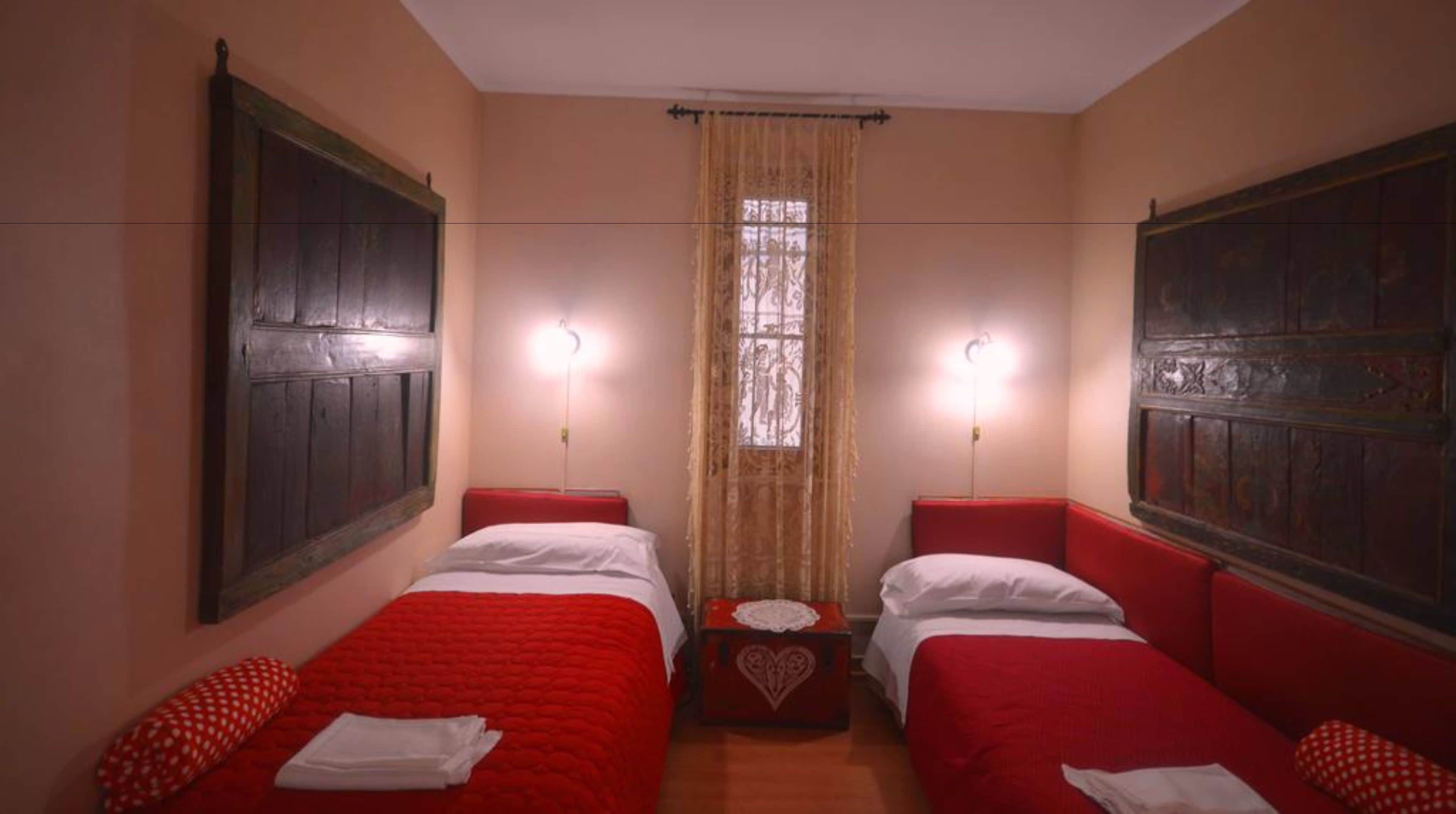 A room with two beds, each with a red headboard and white bedding. There is a small table with a lace tablecloth between the beds.