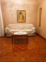 Una habitación con piso de madera, un sofá color beige y una pequeña mesa de madera con mantel de encaje.
