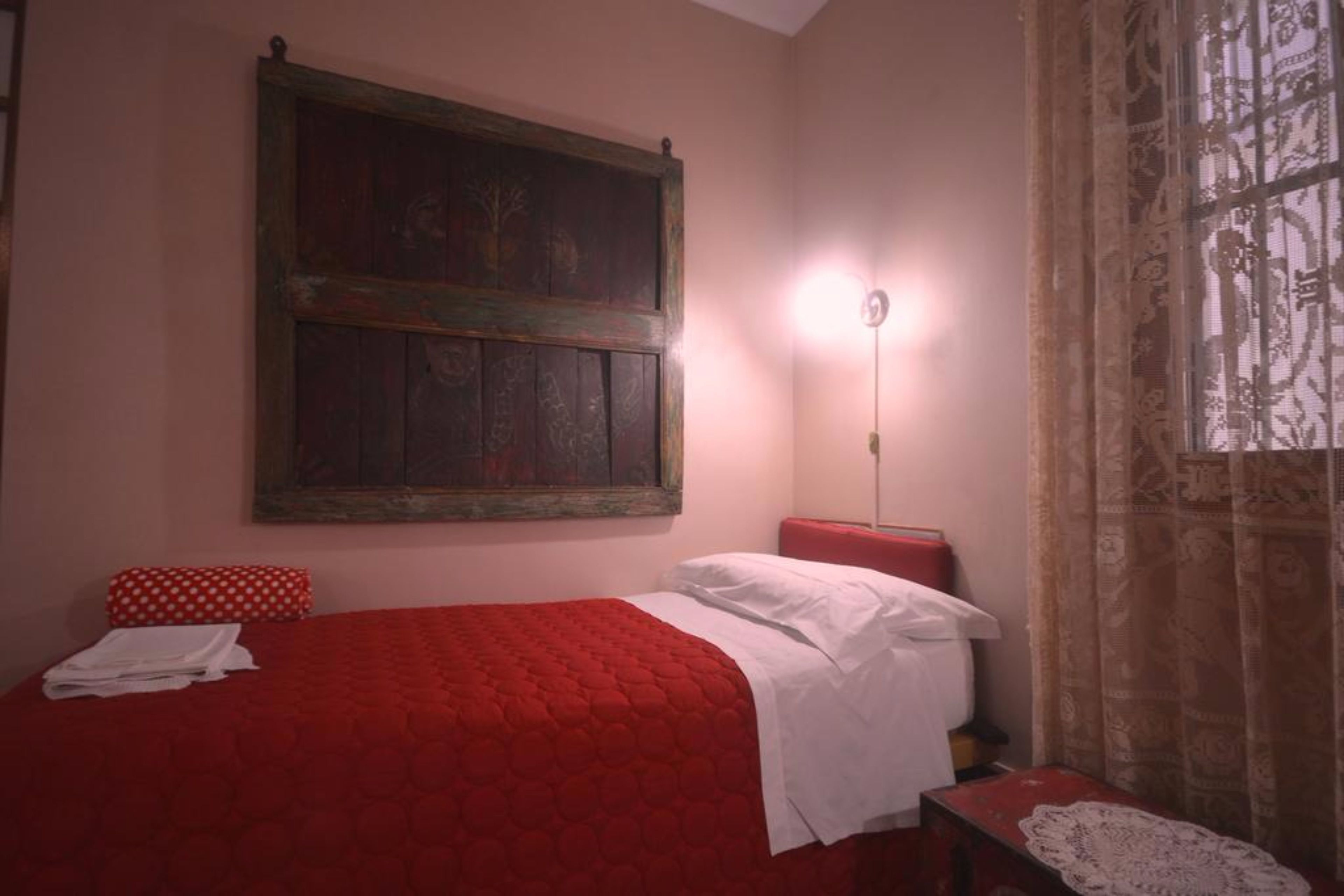 A room with a red bed, white pillows, a red bench, a wooden-framed artwork on the wall, and a patterned curtain.