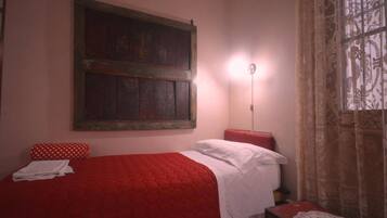 A room with a red bed, white pillows, a red bench, a wooden-framed artwork on the wall, and a patterned curtain.