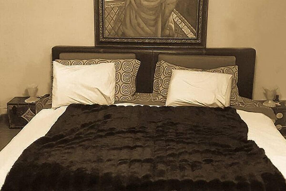 Una cama con una colcha negra, almohadas blancas y ropa de cama estampada. Hay una mesita de noche con una lámpara y un adorno encima.