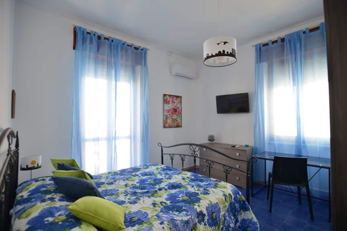 Un dormitorio con una cama, un escritorio y una silla. Hay una ventana con cortinas azules, un cuadro en la pared y un televisor montado sobre el escritorio.