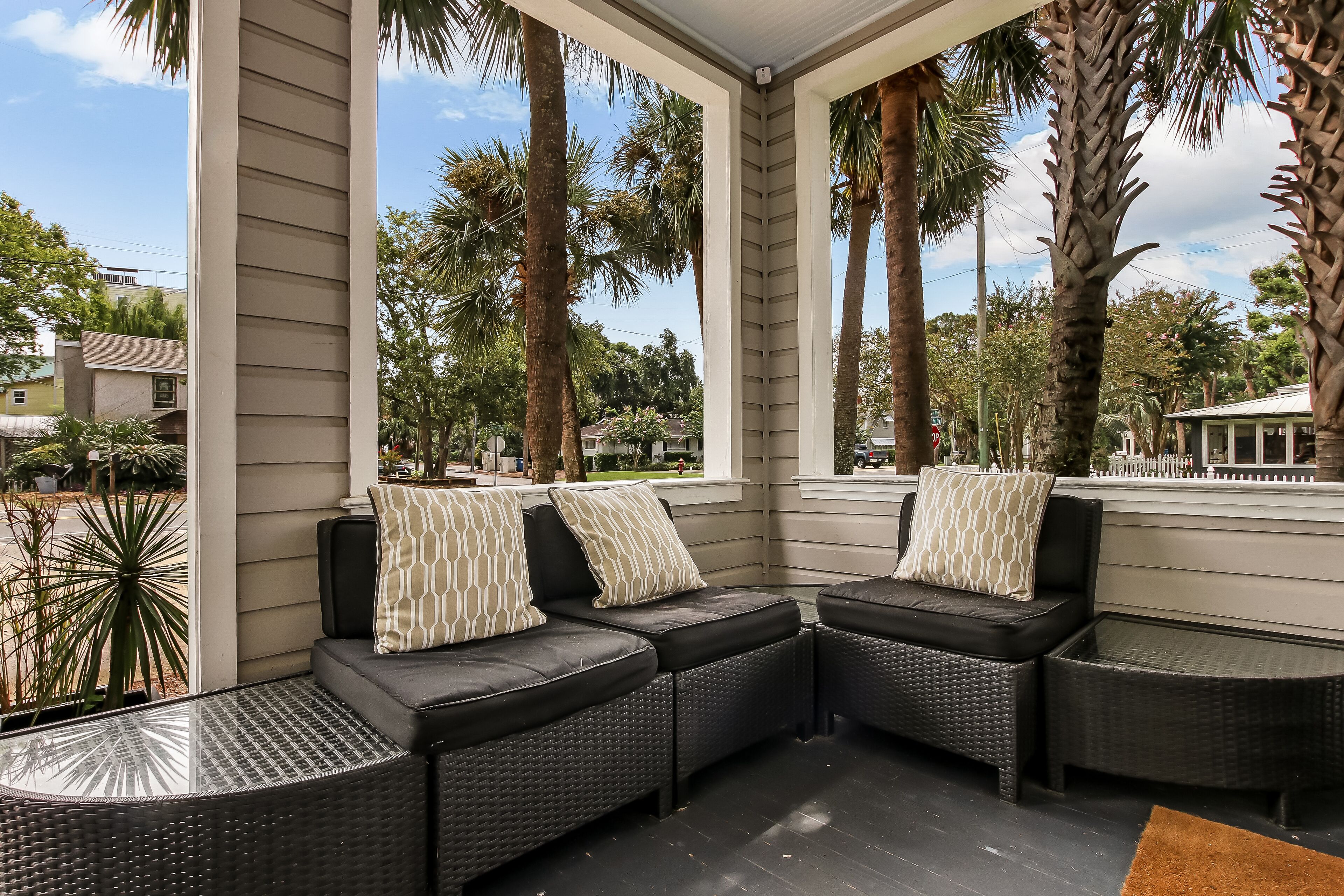 A covered outdoor seating area with a curved black sofa and a glass-top table, surrounded by tall palm trees and a view of the street.