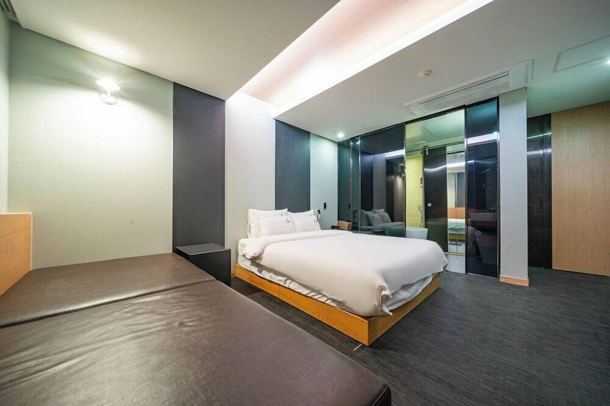 A modern hotel room with a bed, a bench, a mirror, and a sliding door leading to a bathroom.