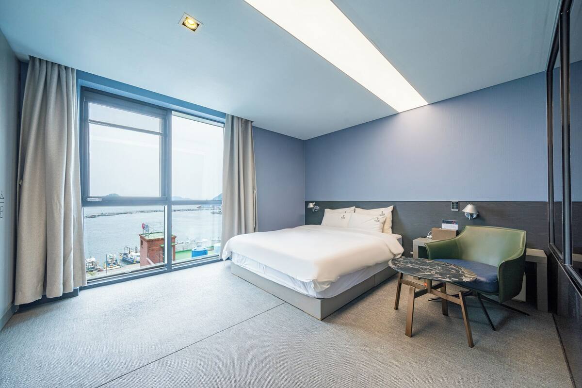 A modern hotel room with a large bed, a desk, a chair, and a view of a body of water.