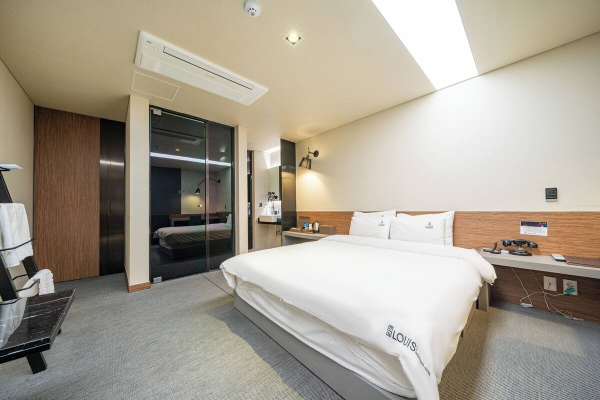 A hotel room with a large bed, a desk, a chair, an air conditioning unit, and a sliding door leading to a bathroom.