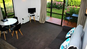 A room with a sofa, a TV, a dining table with chairs, and a view of a garden through large windows.