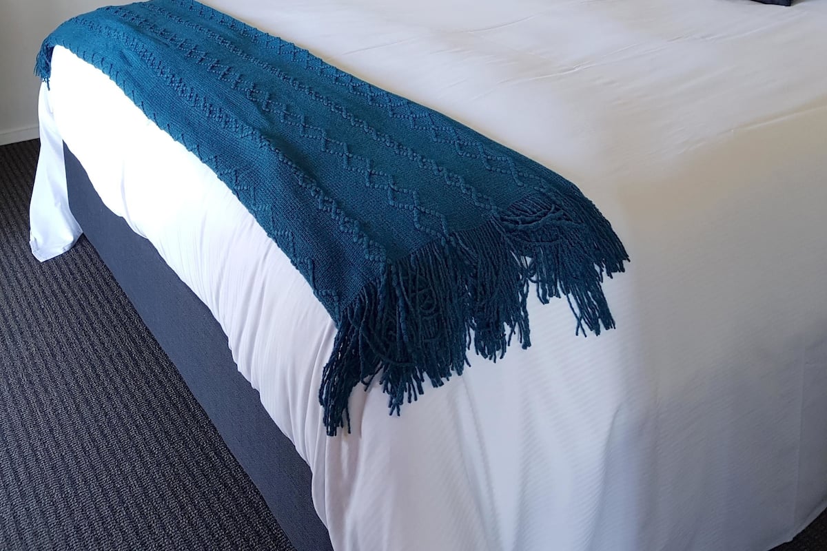 Een netjes opgemaakt bed met een blauwe deken, witte kussens en een ingelijst schilderij aan de muur.