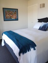 Cama bem arrumada com manta azul, almofadas brancas e um quadro na parede.