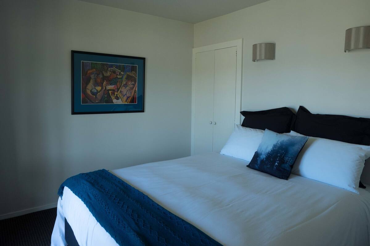 Een net opgemaakt bed met wit en blauw beddengoed, een ingelijst schilderij aan de muur en een gesloten deur.