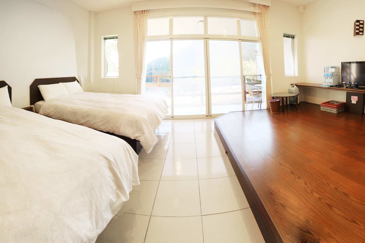 A hotel room with two beds, a wooden floor, a television, and a view of the outdoors through a large window.