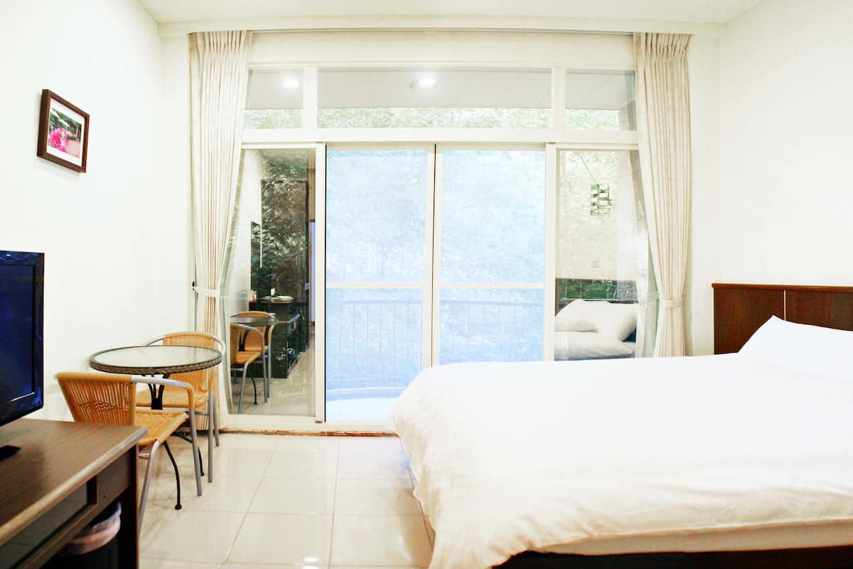 A hotel room with a bed, a TV, a small table with chairs, and a balcony with a view of trees.