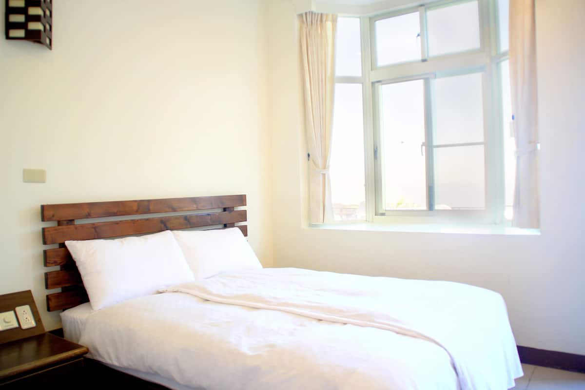 A neatly made bed in a room with a wooden headboard, a window with curtains, and a wall-mounted clock.