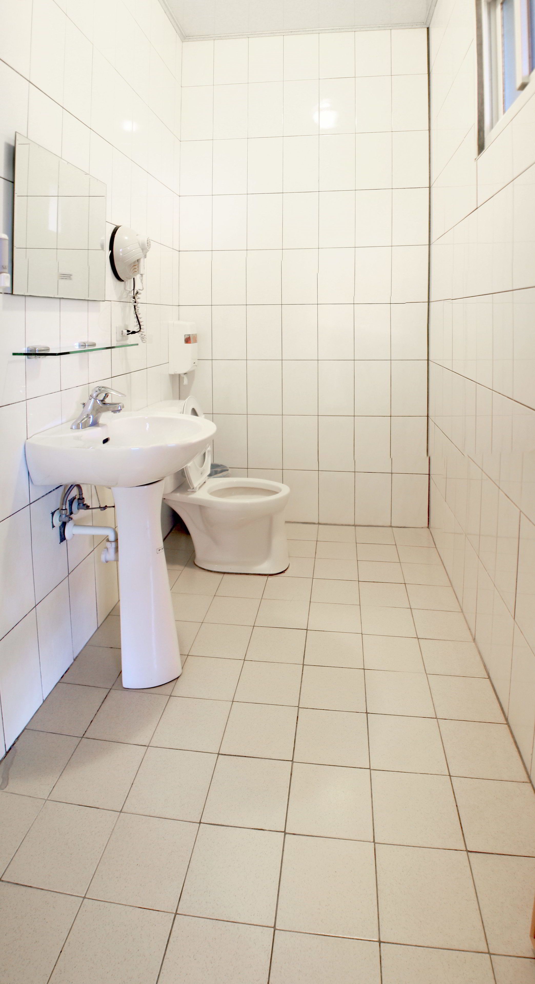 Casa de banho pequena e limpa, com azulejos brancos, lavatório, sanita e secador de cabelo fixado na parede.