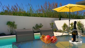 A glass table with a fruit bowl, a bottle, and a glass, near a pool with chairs and an umbrella.