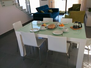 A dining area with a glass table, white chairs, and a blue sofa in the background. - CASA LIMAO (2015) heated pool, near the sea, dunes, forest. (Caldas da Rainha)