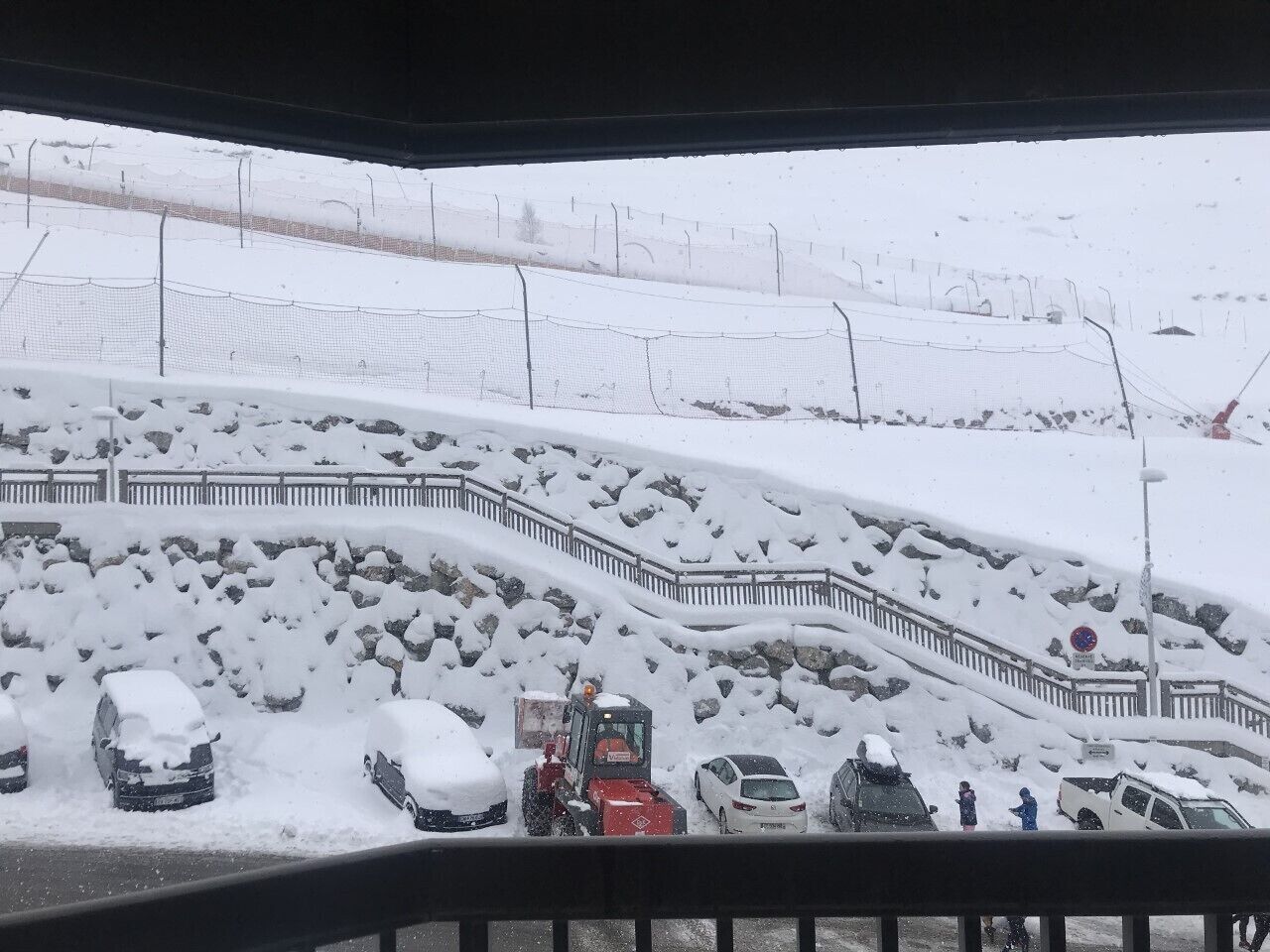 Een besneeerd landschap met een sneeuwbedekte weg, geparkeerde auto's en een sneeuwschuiver.