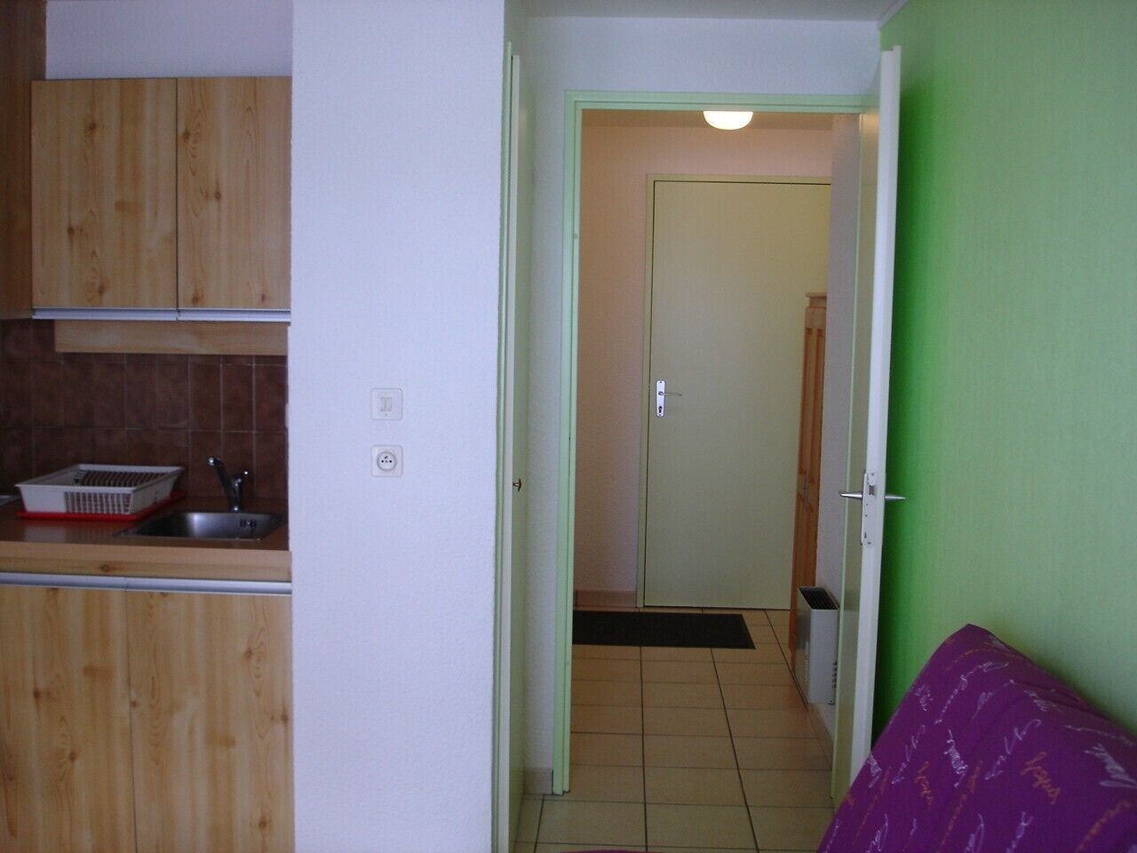 Un couloir étroit avec un coin cuisine sur la gauche, une porte au fond et un canapé violet sur la droite.