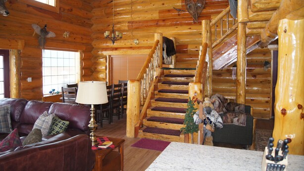 Ein Holzblockhaus-Innenraum mit Ledersofa, Esstisch, Stühlen, einer Treppe und einem Chandelier.