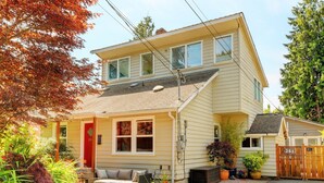 Une maison à deux étages avec une porte rouge, un porche abrité et un petit jardin agrémenté de plantes en pot.