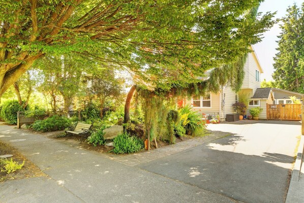 A residential street with a house, a driveway, and a sidewalk lined with trees and shrubs.