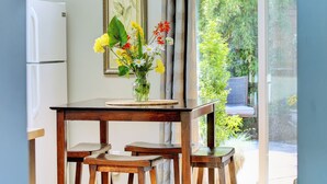 Une table à manger en bois, ornée d’un vase de fleurs, avec un tableau encadré accroché au mur, et une vue sur la verdure à l’extérieur.