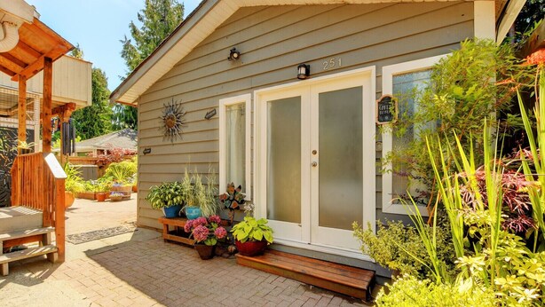 Une maison dotée d’un porche abrité, d’une porte d’entrée avec des panneaux de verre dépoli, et ornée de divers pots de plantes et de fleurs à l’avant.