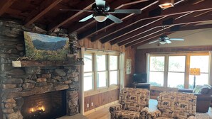 A rustic interior with a stone fireplace, wooden ceiling beams, two patterned armchairs, a wooden coffee table, and a television. - Private Riverfront Cabin: 9 Acres,  Trails to Waterfalls (Sapphire)
