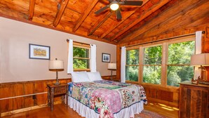 A bedroom with a wooden ceiling, a large bed with a colorful quilt, two bedside lamps, and a view of trees through large windows. - Private Riverfront Cabin: 9 Acres,  Trails to Waterfalls (Sapphire)