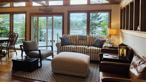 A spacious room with a high ceiling, large windows, a sofa, a chair, a small table, and a lamp. - Oceanfront Maine Acadia Cottage (Hancock)