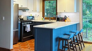 A modern kitchen with wooden beams, a central island, bar stools, and a view of the forest. - Oceanfront Maine Acadia Cottage (Hancock)