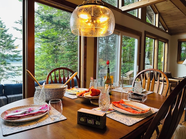 A dining table set with plates, cutlery, and a bottle of wine, with a view of trees through large windows.