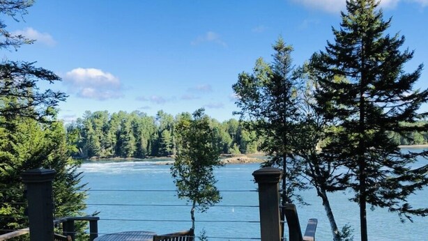 A deck with wooden furniture overlooking a lake, surrounded by trees. - Oceanfront Maine Acadia Cottage (Hancock)