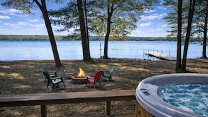 Una terraza con jacuzzi, un fogón y sillas Adirondack junto a un lago.
