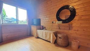 Ein Zimmer mit Holzwänden, einem großen Fenster, einem Fernseher, einem Tisch mit Tischdecke, einem gewebten Korb und einem Mülleimer.