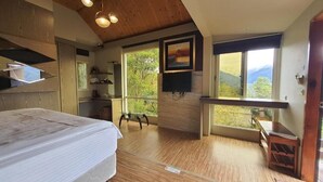 Un dormitorio con una cama grande, techo de madera, televisor montado en la pared y ventana con vistas a la vegetación.