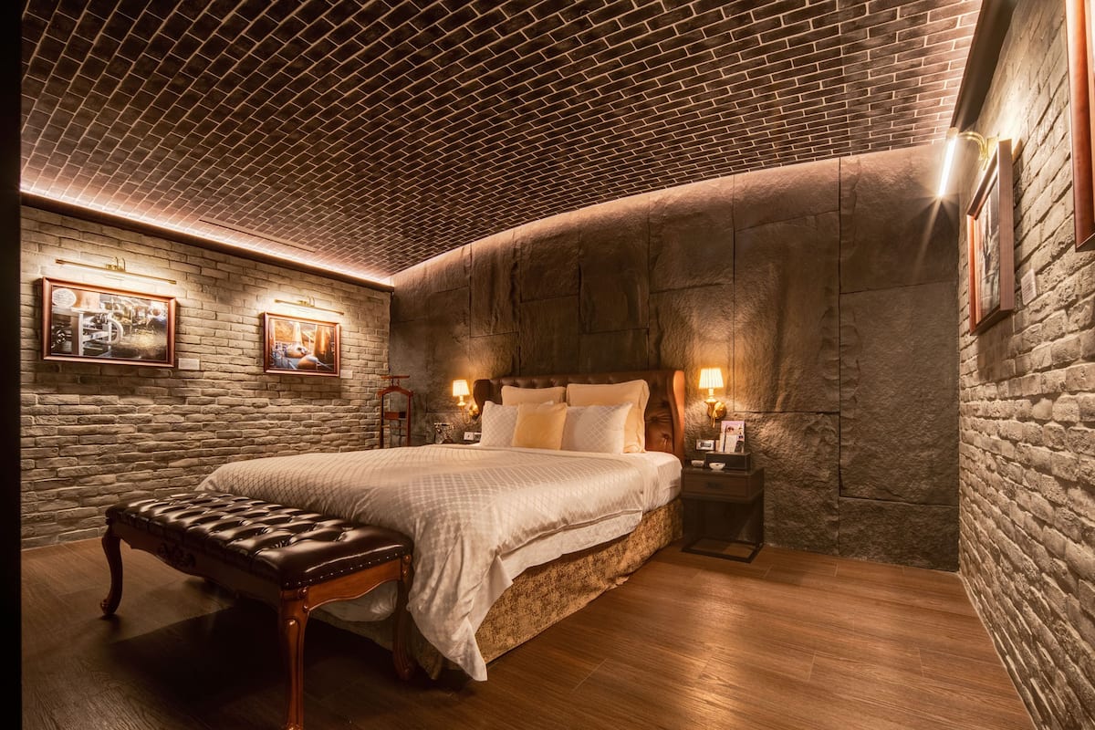 Een slaapkamer met een plafond van baksteenmotief, een groot bed met houten hoofdbord, twee nachtkastjes met lampen en een bankje.